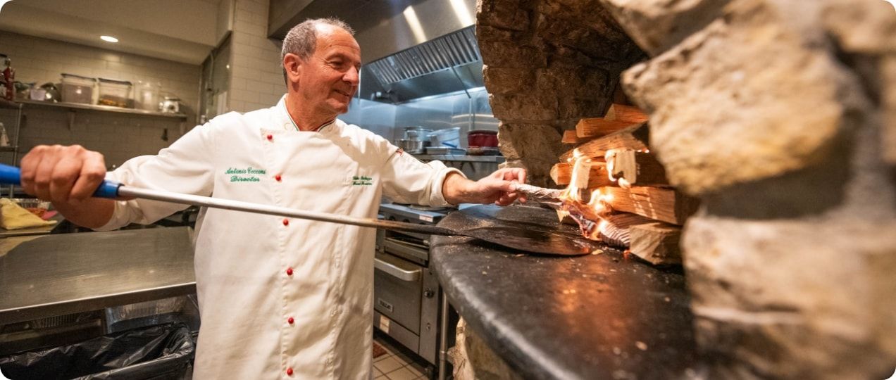 A chef gestures toward the camera while standing in front of a wood oven. 